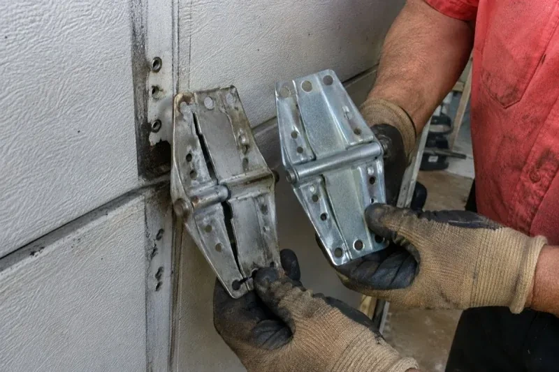 Gloved hands holding two metal garage-door hinges side by side, one discolored with a visible crack and the other bright silver. — Garage Door Hinge Replacement, Fort Erie