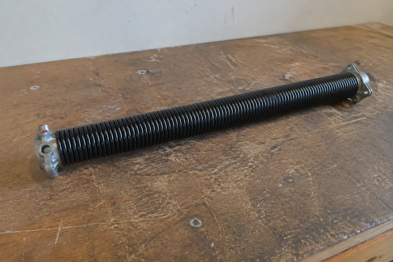 Detached black coil spring with metal end fittings on a workbench. — Torsion Spring Replacement, Fort Erie